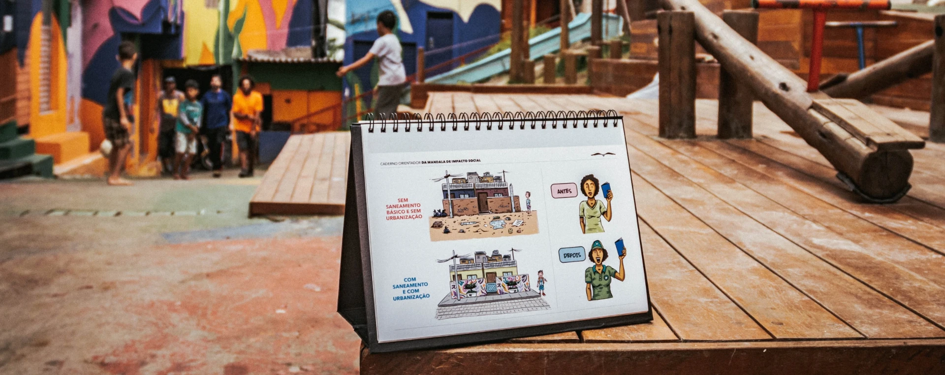 Caderno orientador da Mandala de Impacto Social em primeiro plano, com crianças e a comunidade ao fundo